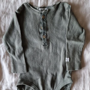 Ribbad grön body från Newbie - Superfin grön ribbad body från Newbie. Långärmad modell med tre knappar framtill för enkel påklädning. Tillverkad i mjuk bomull som är skön mot huden. Någon liten noppra vid knapparna annars inga funna skavanker.