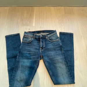 Nudie jeans - Snygga blå jeans från Nudie Jeans Co med klassisk femficksdesign och orangea kontrastsömmar. Jeansen har en rak passform. Bakfickorna har Nudie Jeans signaturbrodyr och läderpatch i midjan.