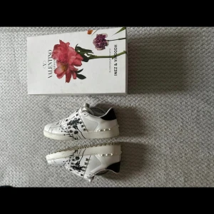 Valentino Garavani Open Stud Flower sneakers - Säljer ett par sjukt snygga Valentino Garavani Open Stud Flower sneakers i vitt skinn med svarta blommiga detaljer och coola nitar på sidorna. Skorna har rund tå, platt sula och svart hälparti. Kommer med originalkartong och extra skosnören.
