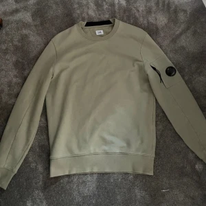 C.P. Company crewneck  - Snygg grön sweatshirt från C.P. Company med rund hals och lång ärm. Tröjan har en ikonisk svart linsdetalj och dragkedjeficka på ärmen. Tillverkad i mjuk bomull för en skön och avslappnad stil. 