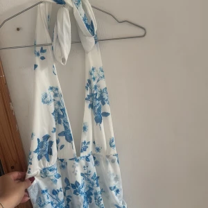 Vit och blå blommig kortklänning - Supersöt kortklänning i vitt med blåa blommor, djup v-ringning och knyt i nacken. Klänningen har en flowy passform och är tillverkad i ett lätt, svalt material som känns perfekt för varma dagar. Volangdetaljer nertill och öppen rygg ger en extra somrig vibe.