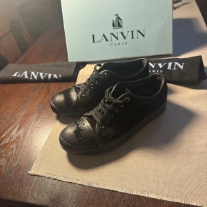 Svarta sneakers från Lanvin Paris - Snygga svarta sneakers från Lanvin Paris med klassisk rund tå och snörning. Skorna har en mix av skinn och textil, med glansiga detaljer på tån och en stilren svart sula. Perfekta för dig som gillar minimalistisk och lyxig streetstyle.