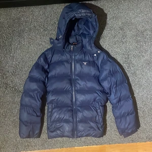 Mörkblå pufferjacka från Gant 146/152 - Snygg mörkblå pufferjacka från Gant med huva och orange innerfoder. Jackan har dragkedja framtill och klassisk Gant-logga på bröstet. Perfekt för kalla dagar och riktigt skön att bära. Passar dig som vill ha en stilren och varm vinterjacka.