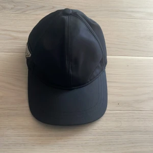 Svart keps från Prada i nylon - Snygg svart keps från Prada med klassisk böjd skärm och ikonisk metalltriangel med logga på sidan. Tillverkad i slät nylon som ger en cool och modern vibe. Perfekt accessoar för att lyfta din streetstyle och ge looken en exklusiv touch.