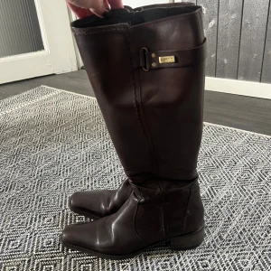 Bruna stövlar i skinn från Esprit - Snygga bruna stövlar från Esprit i äkta skinn med fyrkantig tå och låg klack. Stövlarna har en dekorativ rem med metallspänne upptill och klassisk, stilren design som passar till många outfits. 