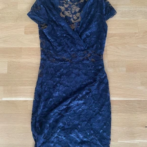 Marinblå spetsklänning Model Behaviour - Snygg marinblå kortklänning från Model Behaviour i storlek S. Klänningen har v-ringad omlottfront, korta ärmar och är helt täckt av blommig spets med transparenta detaljer. Passformen är slim och materialet känns som polyester. Perfekt för dig som vill sticka ut med stil.