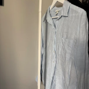 Ljusblå linneskjorta från H&M - Säljer en ljusblå skjorta i linne från H&M. Skjortan har klassisk krage, bröstficka och knäppning framtill. Perfekt för en chill och stilren look, passar till både jeans och shorts. Långärmad och luftig modell.
