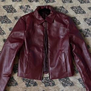 Vinröd / burgundy skinnjacka med dragkedja - Snygg vinröd/burgundy skinnjacka i kortare modell med klassisk krage och raka linjer. Jackan har dragkedja framtill och två fickor på sidorna. Perfekt för dig som vill ha en stilren och edgy look. 😍