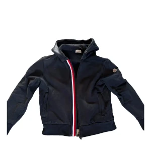 Svart Moncler hoodie zipper - Svart hoodiejacka från Moncler med dragkedja och ikonisk röd, vit och blå rand längs öppningen. Jackan har huva, två fickor och mjukt fleecefoder på insidan. Klassisk Moncler-logga på ärmen och ribbade muddar vid ärmslut och nederkant. TVÅ DRAGKEDJELÄGEN! :)