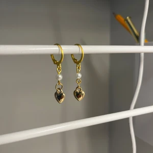 Guldiga örhängen med pärlor och hjärtan - Supersöta örhängen i guldton med små vita pärlor och hängande hjärtan. Perfekt för dig som gillar detaljer och vill ha något som sticker ut lite extra. Hjärtan ger en romantisk vibe och pärlan gör dem extra eleganta. I rostfrittstål men med plast hjärta. Handgjort