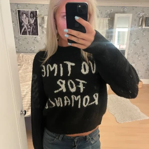 Svart stickad tröja med text - Cool svart stickad tröja med vit text 'NO TIME FOR ROMANCE' framtill. Tröjan har en croppad passform och långa ärmar, perfekt att matcha med jeans. Mjuk och skön känsla, passar dig som gillar statement-plagg och avslappnad stil.