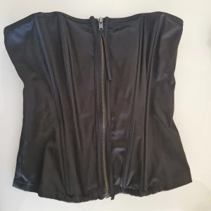 Svart korsett med dragkedja - Snygg svart korsett med markerade sömmar och dragkedja framtill. Den har en glansig finish som ger en cool look. Perfekt för att skapa en edgy outfit till halloween lr utekvällen. Använd 1 gång.