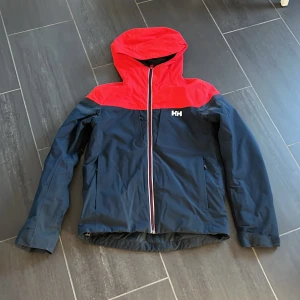 Helly Hansen röd och marinblå vindjacka S - Snygg vindjacka från Helly Hansen i marinblått och rött med huva och hel dragkedja. Jackan har RECCO-reflektor, två sidofickor och liftkortficka. Jackan har en defekt på armen som är lagad, se bild 5. Skick 6/10. Köptes för 1500 kr.