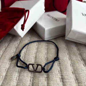 Valentino blå armband  - Supersnyggt armband från Valentino med mörkblå justerbar textilrem och metallfärgad logodetalj i mitten. Armbandet har en minimalistisk design med en liten rund bricka och knytning på sidorna. Perfekt accessoar för dig som gillar stilrena märkesdetaljer.