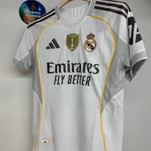 Real Madrid Rodrygo 11 matchtröja - Real Madrid fotbollströja med Rodrygo 11-tryck, Adidas-märke och Champions League 15-patch på ärmen. Vit bas med grå detaljer, svarta ränder och gula linjer. Broderat klubbmärke och sponsortryck framtill. Tillverkad i ventilerande funktionsmaterial.
