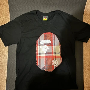 Bape t shirt - Svart t-shirt från BAPE med ikoniskt ape-head tryck i röd- och beigefärgat rutigt mönster både fram och bak. Klassisk passform, korta ärmar och crewneck. Tillverkad i mjuk bomull och har BAPE-logga på ärmen. Perfekt för dig som gillar streetwear vibes.