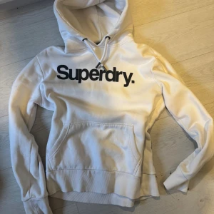 Vit hoodie från Superdry i storlek L - Vit hoodie från Superdry med stor svart logga på bröstet. Klassisk känguruficka framtill, snörning i huvan och ribbade muddar. Mjuk insida och coola Superdry-detaljer på snörändarna. Perfekt för chill dagar eller streetstyle. Sitter som M eller S. Använd bara ett par gånger 
