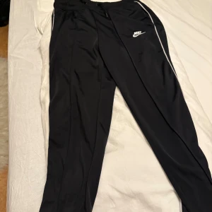 Svarta träningsbyxor från Nike - Svarta mjukisbyxor från Nike med vit logga på låret och vita detaljer längs sidorna. Byxorna har snörning i midjan och är tillverkade i ett mjukt syntetmaterial som passar perfekt för träning. Snygg och sportig look med smal passform.