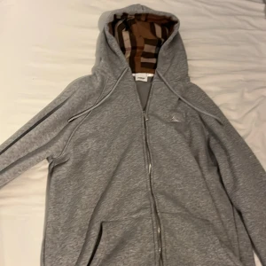 Grå hoodie från Burberry med dragkedja - Grå hoodie från Burberry med klassiskt rutigt foder i huvan. Tröjan har dragkedja framtill, snörning i huvan och två fickor. Storlek L. Testad 1 en gång, Det finns ett hål men den ser man bara från insidan men jag har sytt den 