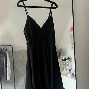 Svart klänning med smala axelband - En stilren svart klänning med v-ringad hals och smala axelband. Klänningen har en lätt utställd kjol och är ärmlös, vilket ger en klassisk och tidlös look. Perfekt för dig som gillar enkel men snygg stil.
