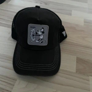 Svart keps med Mickey Mouse-tryck - Cool svart keps med klassiskt Mickey Mouse-motiv framtill. Kepsen har vit kontrastsöm och mesh på sidorna och bak för extra ventilation. Justerbar passform och böjd skärm. Perfekt accessoar för dig som gillar Disney och streetstyle.