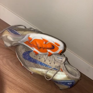 Nike Air Zoom fotbollsskor prototyp - Unika Nike Air Zoom fotbollsskor i Bowerman Prototype edition. Skorna har en skimrande beige ovandel med blå Nike-swoosh och detaljer, samt orange och vit sula med dobbar. Lätta och snabba med snörning, levereras med original vit/blå Bowerman Prototype-påse.