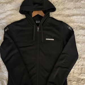 Svart hoodie från EA7 Emporio Armani - Svart hoodie från EA7 Emporio Armani med dragkedja framtill och logga på bröstet och ärmarna. Tröjan har huva med snörning och är tillverkad i ett mjukt material som känns sportigt och stilrent. Perfekt för en chill och snygg look.