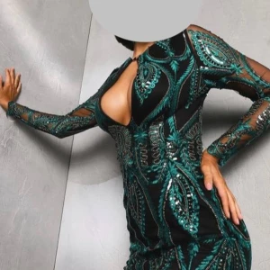 Svart och grön långklänning med slits - Säljer en svart långklänning med grönt paisley-inspirerat mönster och glittriga detaljer. Klänningen har långärmade meshpartier, djup v-ringning med cutout och en snygg slits framtill. Passformen är slim och materialet känns som polyester med paljetter.