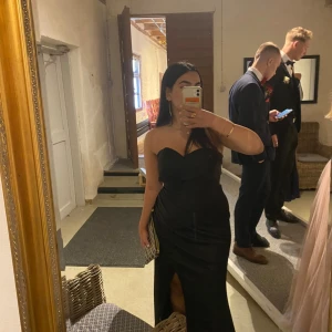 Svart långklänning från Jarlo - Supersnygg svart axelbandslös långklänning från Jarlo med figurnära passform och slits framtill. Klänningen har en elegant drapering och är tillverkad i ett glansigt material som ger en lyxig känsla. Perfekt för bal eller festkvällar! Använd en gång 