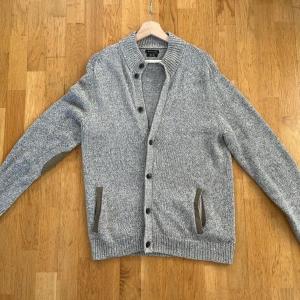 Beige stickad kofta med mockadetaljer - Snygg beigemelerad stickad kofta från massimo Dutti med bruna mockadetaljer på armbågar och vid fickor. Koftan har ribbad krage, muddar och knäppning framtill.