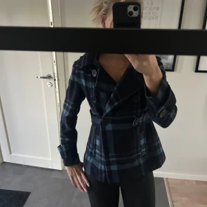 Rutig skepparkavaj  - Snygg skepparkavaj i mörkblått och blått rutigt mönster. Jackan har dubbelknäppning med stora knappar, krage och markerad midja. Finns luva men går att ta bort.