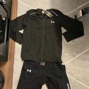 Svart vindjacka från Under Armour - Svart vindjacka från Under Armour med vit logga på bröstet och stor vit text på ryggen. Jackan har hel dragkedja, två sidofickor med dragkedja och diskreta vita detaljer vid axlarna. Perfekt för träning och sportiga dagar.
