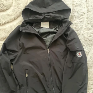 Svart vindjacka från Moncler - Svart vindjacka från Moncler med huva, hel dragkedja och två fickor framtill. Jackan har Moncler-logga på ärmen och elastiska muddar vid ärmslut och midja. Tillverkad i lätt och slitstark polyester, perfekt för blåsiga dagar.