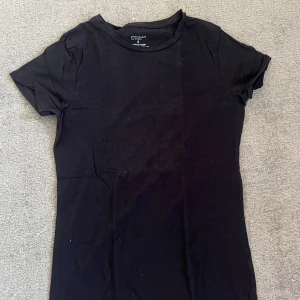 Svart stretchig topp från Primark - En enkel och stilren svart topp från Primark med rund hals och korta ärmar. Modellen är stretchig och sitter tight mot kroppen, perfekt för att matcha med jeans eller kjol. Materialet är mjukt och bekvämt bomull med lite stretch.