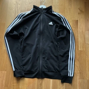 Svart Adidas track jacket med vita ränder - Svart Adidas track jacket med klassiska vita ränder längs ärmarna och vit logga på bröstet. Jackan har hel dragkedja, hög krage och ribbade muddar. Tillverkad i polyester för en sportig och bekväm känsla.