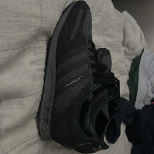 Svarta Adidas LA Trainer sneakers - Säljer ett par svarta Adidas LA Trainer sneakers med klassiska tre ränder på sidan och snörning. Skorna har en platt sula med gummidubbar för extra grepp och är tillverkade i syntetmaterial. Perfekt för dig som gillar sportig och stilren look.