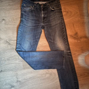 Nudie Jeans blå slim fit jeans 31/34 - Säljer ett par klassiska blå jeans från Nudie Jeans i modellen slim fit. Jeansen har fem fickor, snygga slitningar och Nudie-loggan på bakfickan. De är tillverkade i mjukt denimtyg och har en normal passform med raka ben.