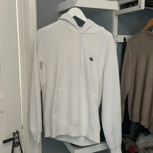 Vit hoodie från Lacoste med huva - Säljer en stilren vit hoodie från Lacoste i klassisk passform. Tröjan har huva med dragsko, magficka och den ikoniska krokodilloggan på bröstet. Perfekt för dig som gillar enkel och clean stil.