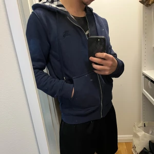 Marinblå Burberry hoodie med dragkedja - Säljer en marinblå hoodie från Burberry med klassiskt rutigt foder i huvan. Tröjan har dragkedja, snörning i huvan och två fickor framtill. Perfekt för dig som vill ha en stilren och lyxig look med streetkänsla. Helt oanvänd i nytt skick st S