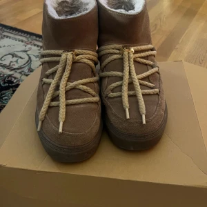 Beige Inuikii boots med snörning - Säljer ett par beige boots från Inuikii i mocka med grova snören och guldiga detaljer. Skorna har rund tå, platt sula och är fodrade med mjuk päls för extra värme. Perfekta för kalla dagar och har en snygg, chunky look.