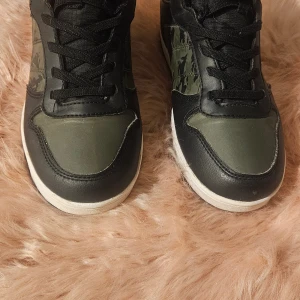 Svarta och gröna sneakers med camomönster - Snygga fodrade sneakers i svart och olivgrönt med coolt camouflagemönster på sidan. Skorna har snörning, kardborreband och vit platt sula. Materialet är syntet och skinnimitation, och de är märkta med 'Water Point'. Perfekta för dig som gillar streetstyle.