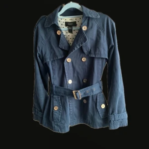 Mörkblå trenchcoat från Mango - Snygg mörkblå trenchcoat från Mango med klassisk dubbelknäppning, bälte i midjan och dekorativa knappar. Jackan har långa ärmar, axelklaffar och ett prickigt innerfoder som ger en cool kontrast. Perfekt för dig som vill ha en stilren och tidlös look.