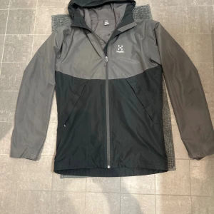 Svart och grå vindjacka från Haglöfs - Snygg svart och grå vindjacka från Haglöfs med huva och hel dragkedja framtill. Jackan har två sidofickor med dragkedja och justerbar dragsko i huvan. Perfekt för blåsiga dagar och outdoor-aktiviteter. Klassisk logga på bröstet och stilren design.