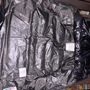 Svart Moncler dunjacka med stor logga - Snygg svart dunjacka från Moncler med glansig finish och stor vit broderad logga på ärmen. Jackan har dragkedja och klassisk Moncler badge. Insidan har serietidningsmönstrad tvättrådslapp och märkning för äkta dun. Perfekt för kalla dagar.