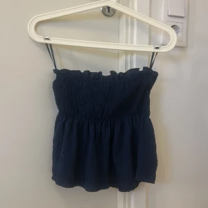 Mörkblå smockad linnetopp - Supersöt mörkblå linnetopp med smockad överdel och volangkant upptill. Tubtoppen en luftig, lätt rynkad nederdel som ger en avslappnad och trendig look. Perfekt för varma dagar och enkel att matcha med jeans eller kjol.