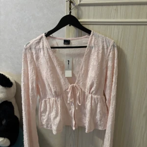 Ljusrosa blus med knyt från Gina Tricot - Supersöt ljusrosa blus från Gina Tricot med v-ringning och knyt framtill. Blusen har lång ärm och är gjord i ett tunt, broderat tyg med struktur. Den har en peplum-liknande form som ger en feminin siluett. Perfekt för dig som gillar romantiska detaljer!