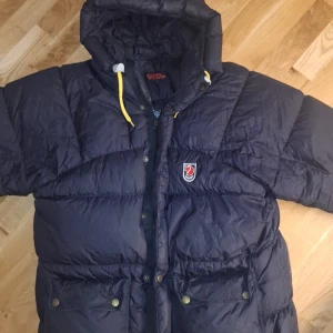 Svart dunjacka från Fjällräven XS - Säljer en svart dunjacka från Fjällräven i storlek XS. Jackan har huva med gula snören, dragkedja och tryckknappar framtill samt två stora fickor med lock. Klassisk Fjällräven-logga på bröstet och quiltad design som håller värmen under kalla dagar. Den har en liten defekt på högra armen men  i övrigt är den i fint skick. Pris kan diskuteras.
