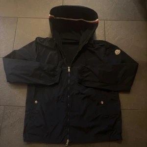 Moncler windbraker - Svart vindjacka från Moncler med klassisk logga på ärmen, hel dragkedja framtill och huva med röd och vit rand på insidan. Jackan har elastiska muddar vid ärmsluten och två fickor med knappstängning. Perfekt för dig som vill ha en stilren och sportig look. Nypris 9000 men får den för 3499 och 3 i Moncler stolek