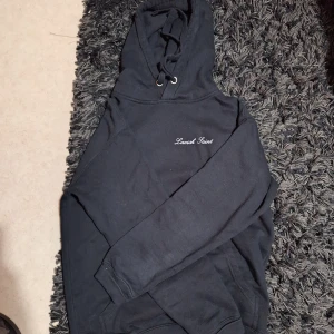 Svart hoodie Lavish Saint med tryck - Svart hoodie från Lavish Saint med broderad logga på bröstet och stort tryck på ryggen med texten 'Money talks. We translate.' och tidningsmotiv. Klassisk huva med snörning och långa ärmar. Perfekt för dig som gillar streetwear vibes.