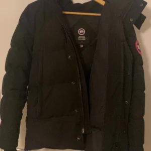 Svart dunjacka från Canada Goose M - Säljer en svart dunjacka från Canada Goose i storlek M. Jackan har klassisk pufferstil, hög krage och Canada Goose-logga på ärmen. Fylld med dun för extra värme och har dragkedja framtill. Perfekt för kalla vinterdagar. Pris kan diskuteras. 